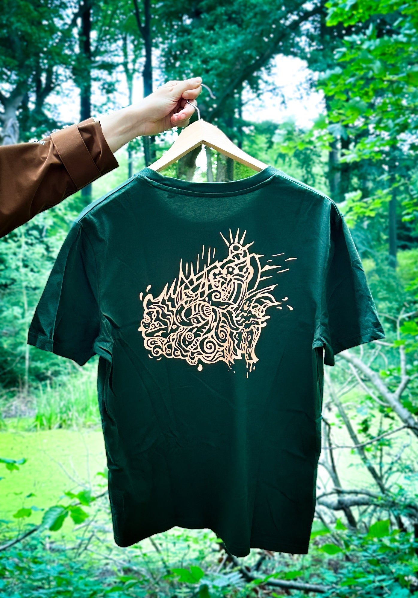 Green SOAL Studio t-shirt back with a orange art Seawave print on a hanger against a forest background