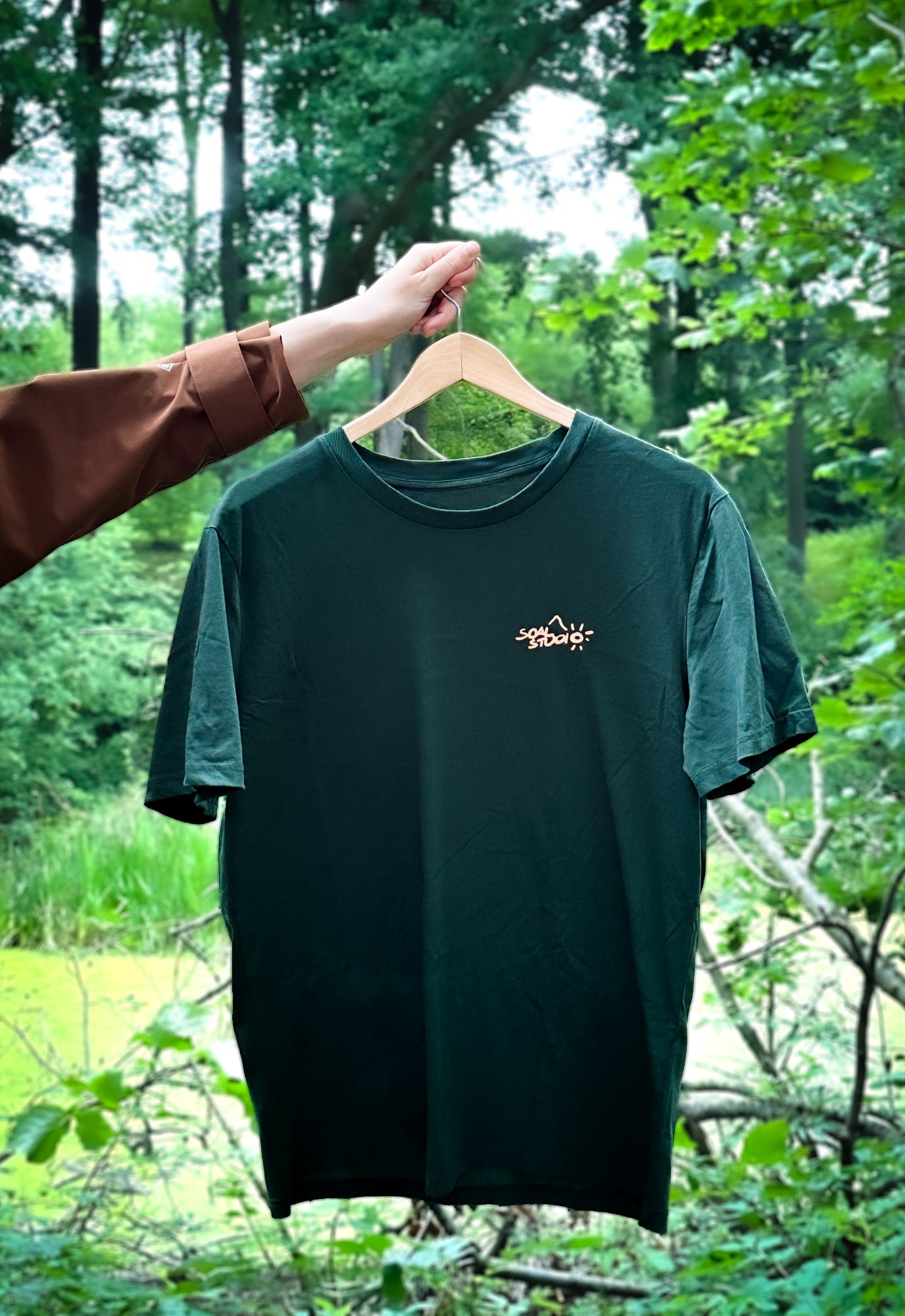 Green t-shirt with a orange SOAL Studio logo on a hanger against a forest background