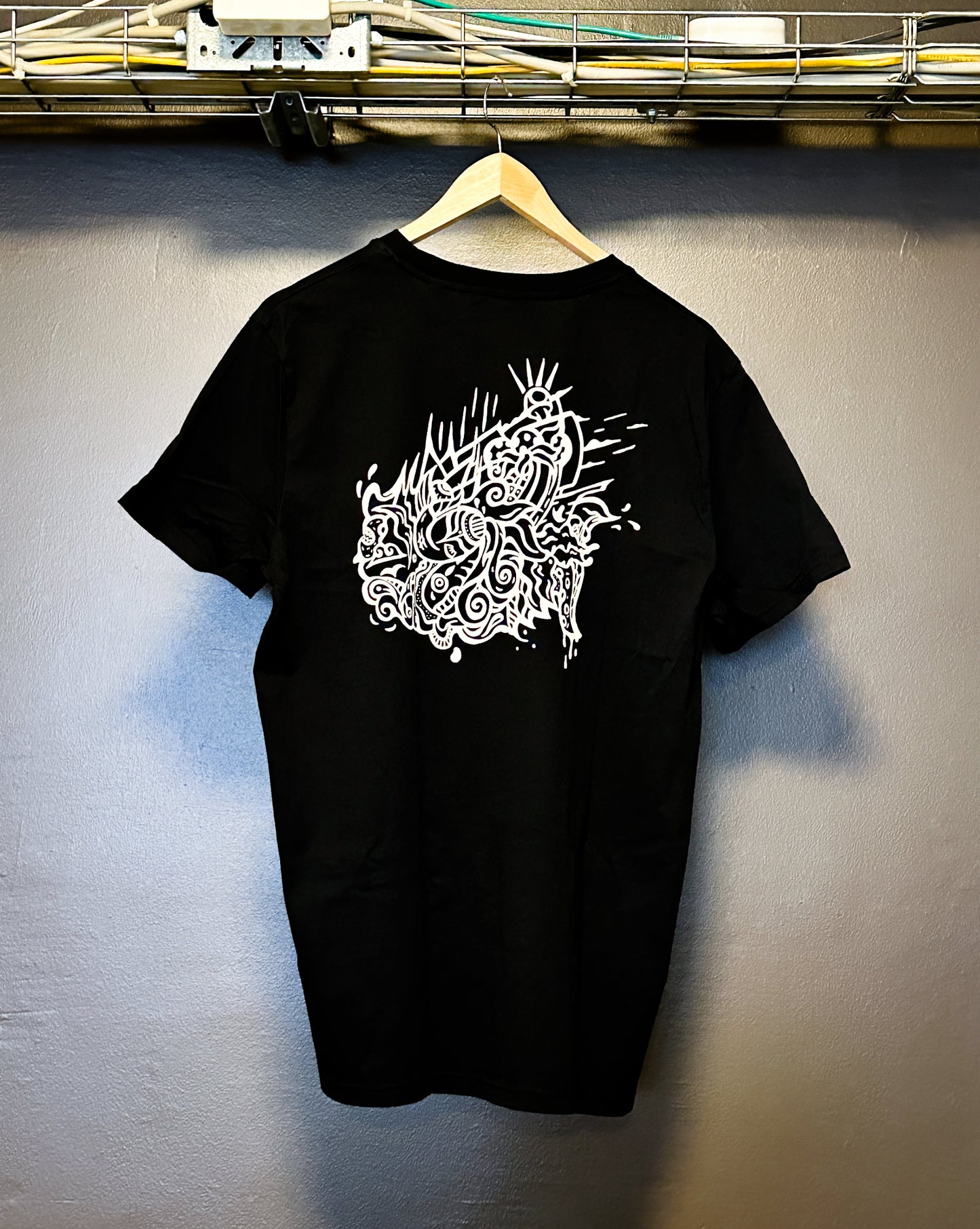 Black SOAL Studio t-shirt back with off-white art graphic Seawave on a hanger against a gray wall.