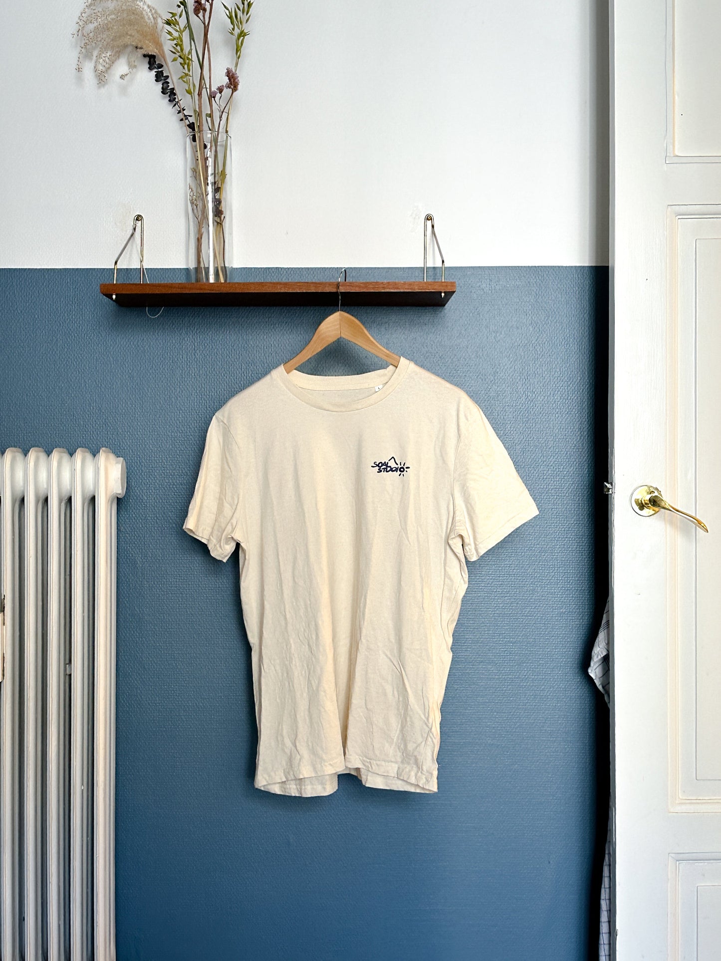 Beige SOAL Studio t-shirt with a SOAL Navy logo on a hanger against a blue wall.