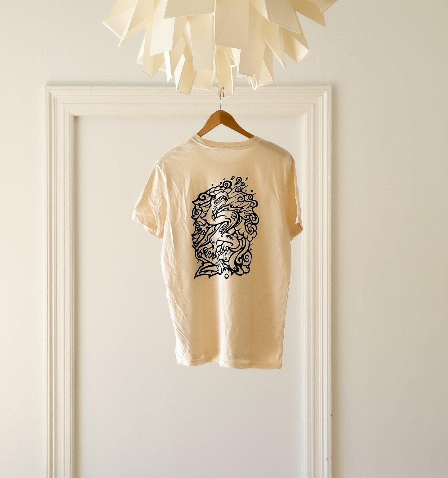 Beige SOAL Studio t-shirt back with navy art graphic Dragon hanging on a wooden hanger against a white ballroom wall.