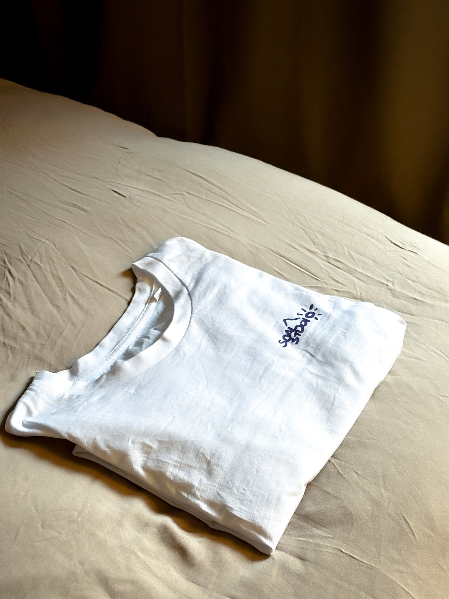 White t-shirt with a purple SOAL logo on a beige bed sheet