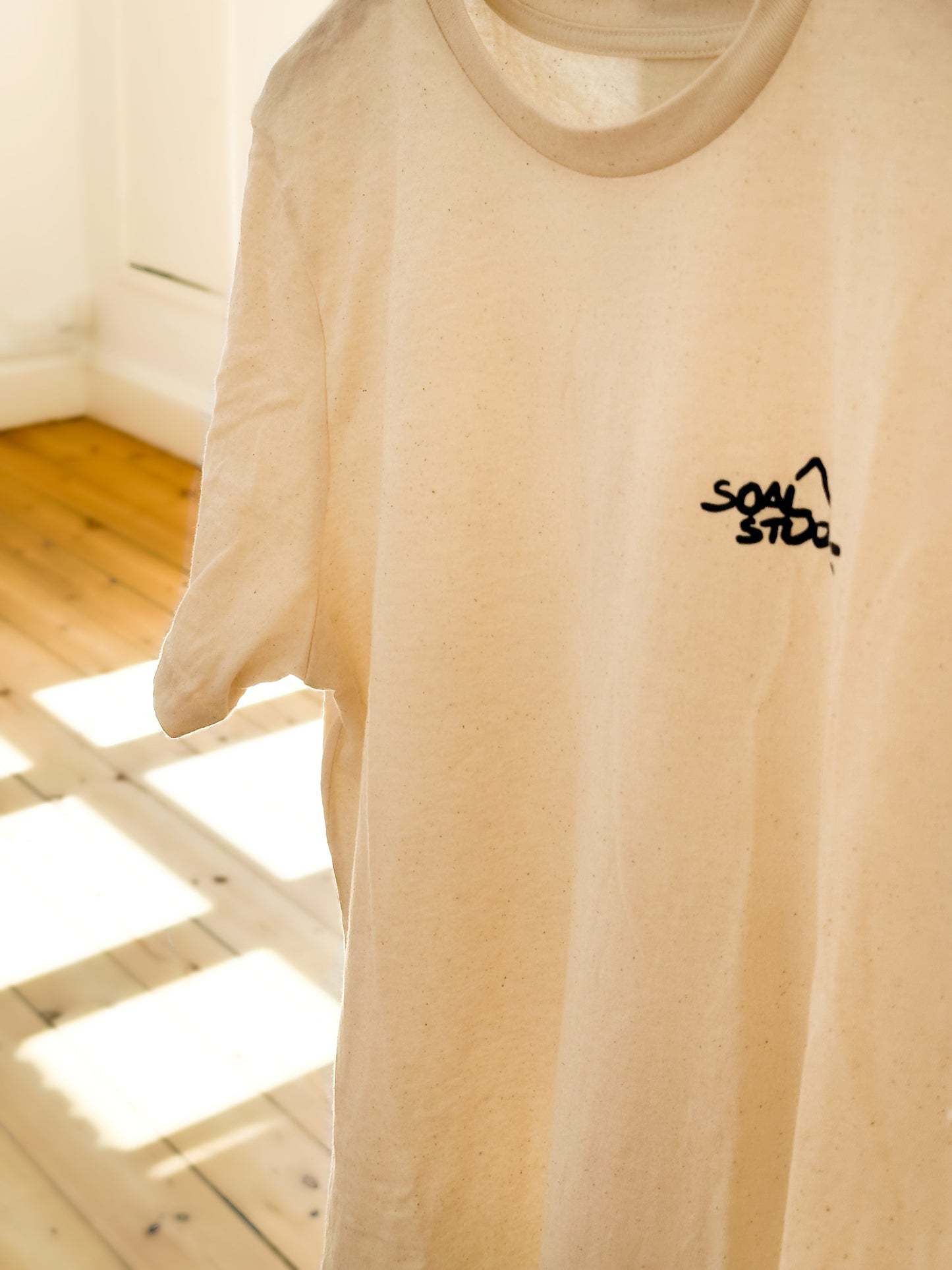 Beige SOAL Studio t-shirt front with navy art graphic navy logo t-shirt with a navy SOAL logo on a wooden floor