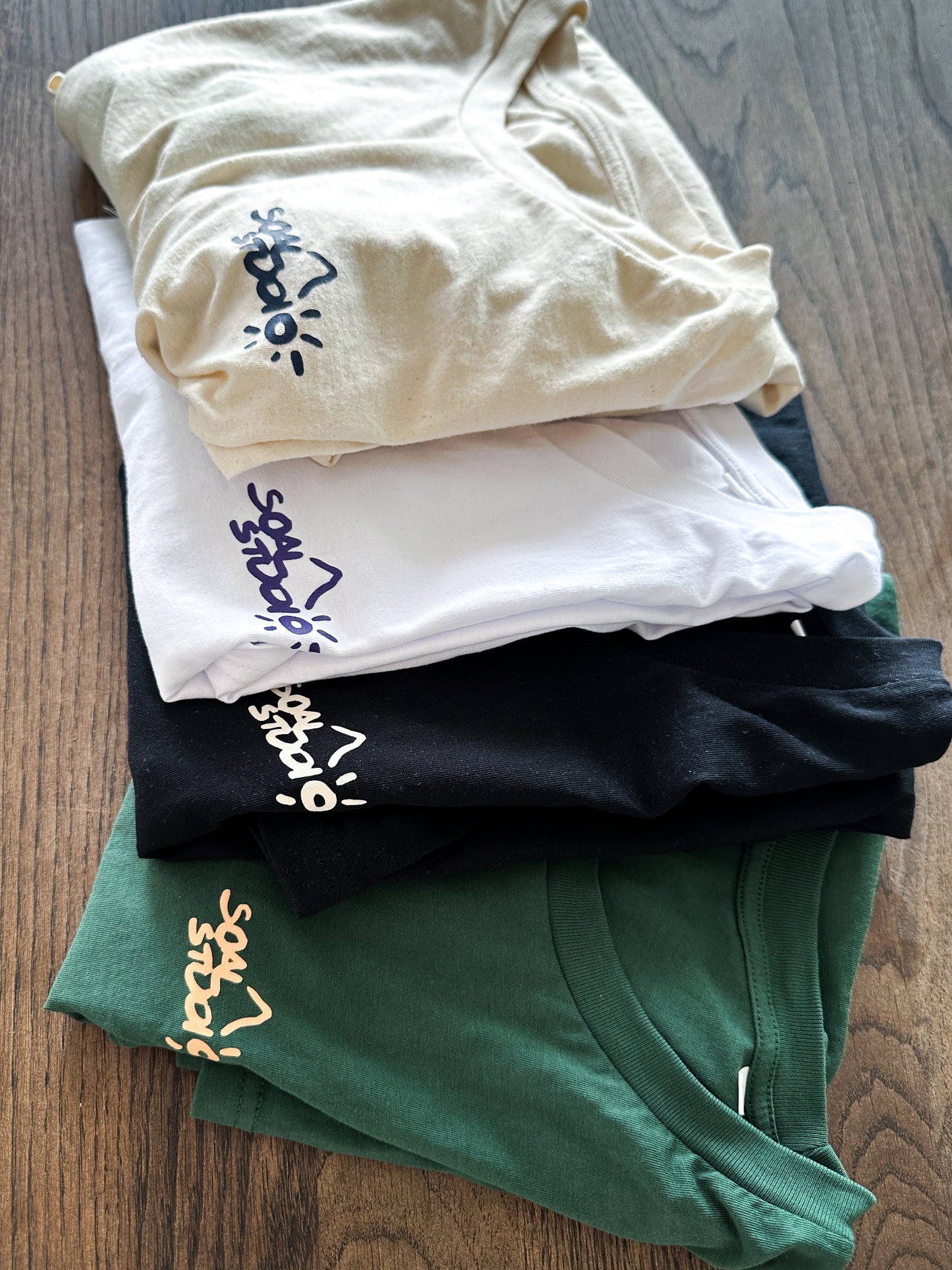 All Editions of SOAL Studio art-shirts Batch #1 - Stack of folded t-shirts in beige, white, black, and green on a wooden surface.
