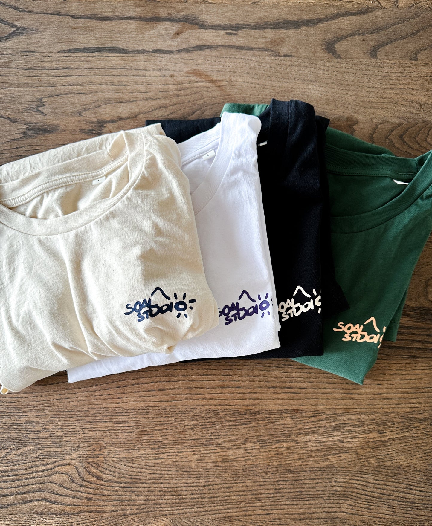 Four SOAL Studio folded t-shirts in beige, white, black, and green on a wooden surface. This is all the editions from the first batch.