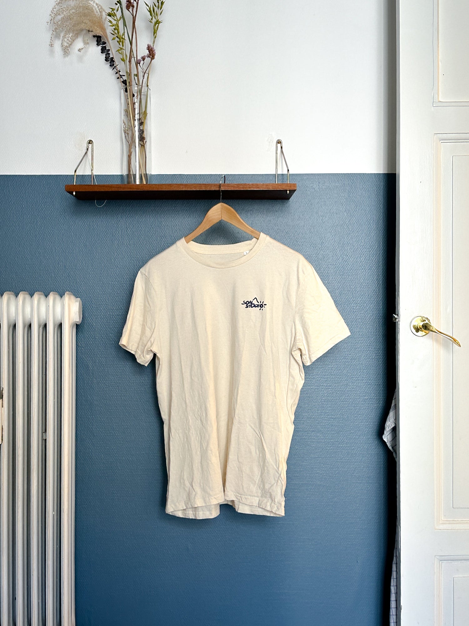 Beige SOAL Studio t-shirt with a SOAL Navy logo on a hanger against a blue wall.