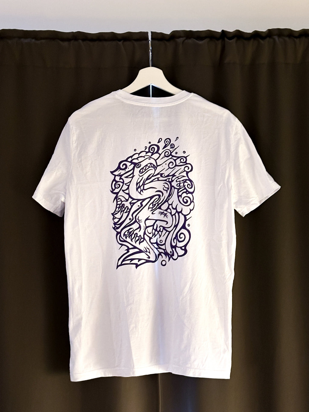 White SOAL Studio t-shirt with a purple dragon design on a hanger against a dark curtain background